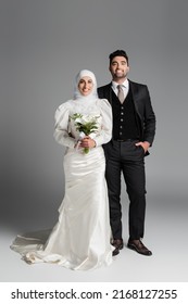 cheerful groom in suit posing near muslim bride with wedding bouquet on grey