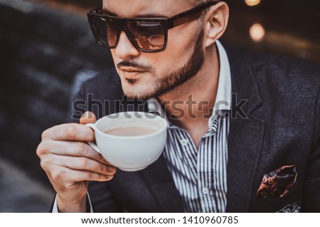 Cheerful groomed gentelman in sunglasses is enjoying his coffeebreak with cup of fresh latte.