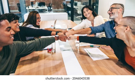 Cheerful group of businesspeople bringing their fists together while sitting in a boardroom. Diverse group of colleagues smiling happily during a meeting in modern workplace. - Powered by Shutterstock