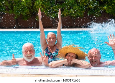 Cheerful Group Of Four Senior People Floating In Outdoor Swimming Pool Raising Splashes Of Water. They Smile Relaxed On Vacation Under The Bright Sun