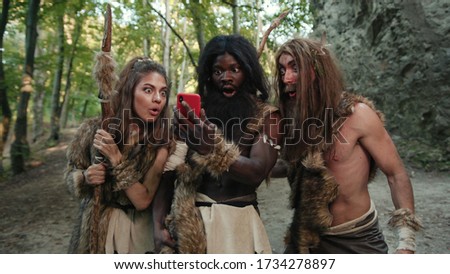 Cheerful group of neanderthal hunters using modern technology smartphone cheering up walking outside cave in the jungle. Technology and first people.