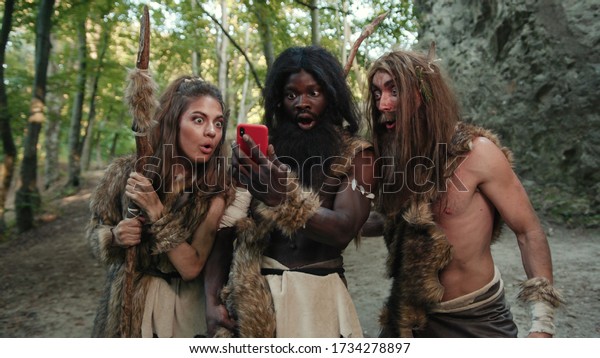 Cheerful group of neanderthal hunters using modern technology smartphone cheering up walking outside cave in the jungle. Technology and first people.