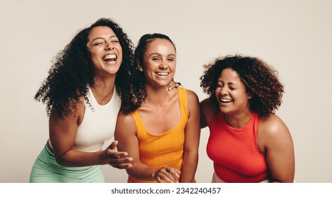 Cheerful group of three young, diverse female athletes celebrate their friendship and healthy lifestyle while wearing fitness clothing in a studio. Sporty young women laughing together. - Powered by Shutterstock