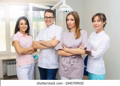 Cheerful Group Of Young Dentists And Their Assistants Standing In The Dental Office And Looking At Camera And Friendly Smiling At White Background Of Medical Room.