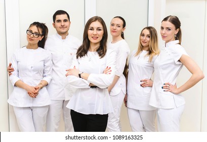 Cheerful Group Of Young Dentists And Their Assistants Standing In The Dental Office And Looking At Camera And Friendly Smiling At White Background Of Medical Room.