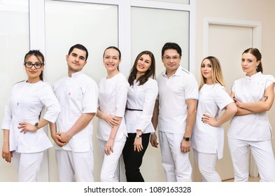 Cheerful Group Of Young Dentists And Their Assistants Standing In The Dental Office And Looking At Camera And Friendly Smiling At White Background Of Medical Room.