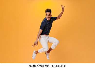 Cheerful Guy In Black T-shirt Jumping High On Orange Background. Snapshot Of Man In White Pants Dancing And Having Fun On Isolated