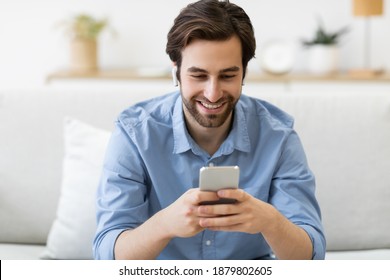 Cheerful Guy Making Video Call Using Smartphone And Earbuds Sitting On Sofa At Home. Man Playing Mobile Game In Living Room Indoors. Gadget, New Application Concept