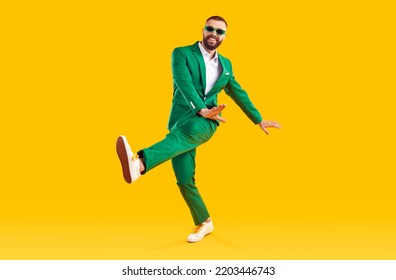 스튜디오에서 춤을 추는 세련된 파티 의상을 입은 쾌활한 남자. 밝은 노란색의 배경을 배경으로 춤을 추는 유행의 녹색 정장과 선글라스를 입은 행복한 남자의 전신 초상화 스톡 사진