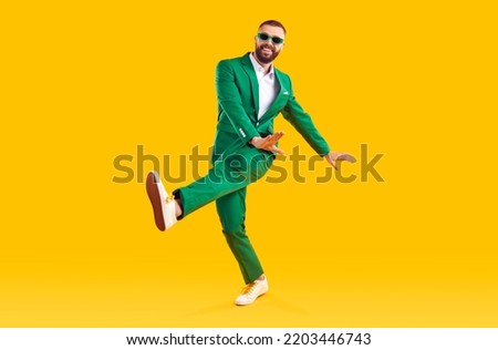 Cheerful guy in a stylish party outfit dancing in the studio. Full length portrait of a happy man wearing a fashionable green suit and sunglasses dancing isolated on a bright yellow color background