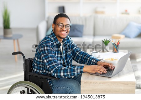 Cheerful handicapped black man in wheelchair using laptop for online job from home, having business meeting on web. African American disabled freelancer working on remote project