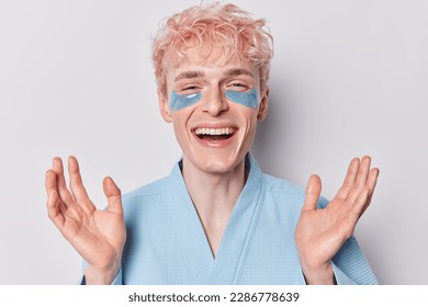 Cheerful handsome adult man with pink hair spreads palms smiles broadly has happy mood applies blue hydrogel patches under eyes to reduce wrinkles and puffiness dressed in domestic bathrobe. - Powered by Shutterstock