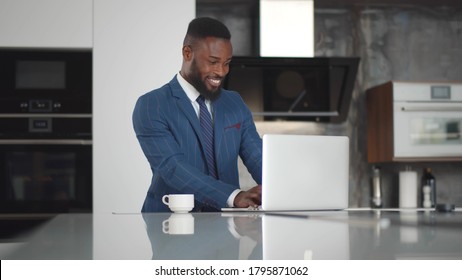 Cheerful handsome african businessman wearing suit working with laptop at modern kitchen. Afro-american entrepreneur checking email on computer during breakfast in kitchen at home - Powered by Shutterstock