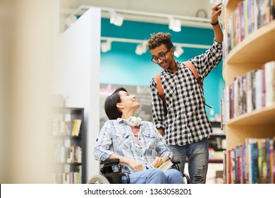 Cheerful handsome black guy with Afro hairstyle helping disabled student girl to take book from shelf in library
