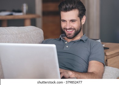 Cheerful And Handsome Blogger. Cheerful Handsome Young Man Using His Laptop With Smile While Sitting On The Couch At Home