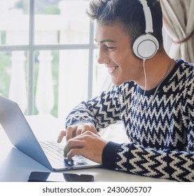 Cheerful handsome boy using laptop at home in front of the window wearing headphones. Zoomer generation boy enjoying technology and social - Powered by Shutterstock