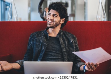 Cheerful handsome ethnic man sitting on red sofa in cafe holding laptop and printed picture and smiling away happy with result of work - Powered by Shutterstock