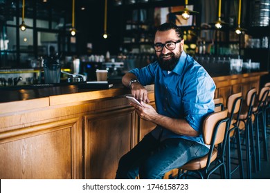 Cheerful Handsome Mature Man In Casual Wear And Spectacles Sitting Near Bar Indoors With Smartphone, Happy Prosperous 40 Years Male Owner Of Small Business Satisfied With Income From Cafetaria