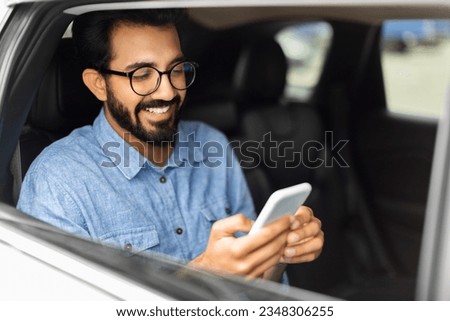 Cheerful handsome millennial eastern guy in casual wearing eyeglasses sitting on auto back seat, using smartphone and smiling, indian man chatting with friends while sitting in taxi