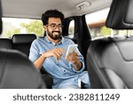 Cheerful handsome millennial indian guy in casual wearing eyeglasses sitting on auto back seat, using smartphone and smiling, hindu man chatting with friends while sitting in taxi