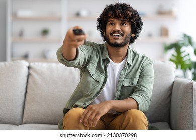 Cheerful Handsome Millennial Indian Guy In Casual Sitting On Couch At Home, Watching Movie, Holding Remote Controller From TV And Smiling, Enjoying His Lazy Weekend, Panorama With Copy Space