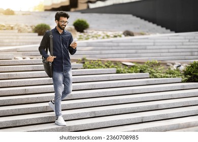 Cheerful handsome millennial indian guy employee going down the stairs outdoors, using cell phone on street, copy space. Happy young eastern man checking email, scrolling on smartphone, full length - Powered by Shutterstock