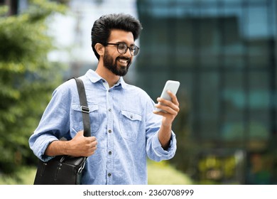 Cheerful handsome millennial indian guy employee going to office in the morning, using cell phone on the street, copy space. Happy young eastern man checking email, scrolling on smartphone outdoors - Powered by Shutterstock