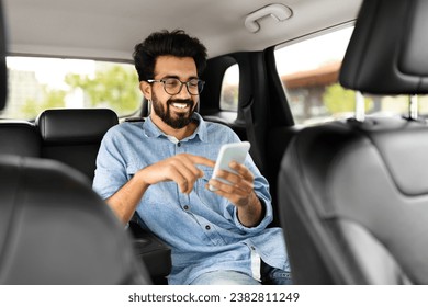 Cheerful handsome millennial indian guy in casual wearing eyeglasses sitting on auto back seat, using smartphone and smiling, hindu man chatting with friends while sitting in taxi