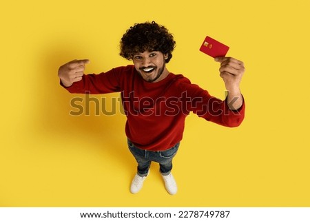 Cheerful handsome millennial indian man in casual pointing at red plastic bank card and smiling, recommending contactless payment, easy fast banking, yellow studio background, top view, copy space