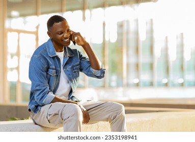 Cheerful handsome stylish young black guy sitting on parapet on street, talking on phone with mate or girlfriend and smiling, copy space. Communication and gadgets, connection concept - Powered by Shutterstock