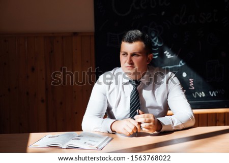 Cheerful handsome teen boy sitting at his desk with book. Emotional portrait of  happy cute smiling male. Lucky guy in white shirt celebrating triumph. Back to school and education concept