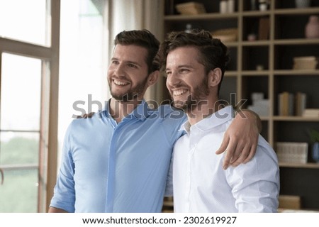 Cheerful handsome young adult twin brothers posing for shooting together, standing close, hugging, patting shoulders, looking away, smiling, laughing, enjoying friendship
