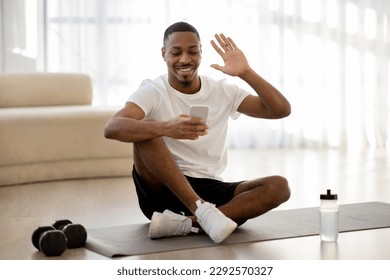 Cheerful handsome young african american guy sportsman sitting on yoga mat, waving at smartphone screen and smiling, greeting fitness coach, have online lesson, home interior, copy space - Powered by Shutterstock