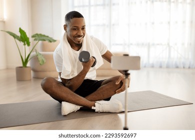 Cheerful handsome young black man in sportswear sitting on yoga mat, training with dumbbell, smiling at smartphone camera, fitness coach recording video at home, copy space. Online workout - Powered by Shutterstock