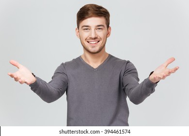 Cheerful handsome young man in grey pullover smiling and showing welcoming gesture over white background