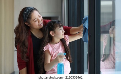 Cheerful And Happy Asian Woman Smiling While Washing Or Cleaning Window Glass Surface With Rag And Spray Detergent. Single Parent Mon And Little Girl Doing House Cleaning Job Concept