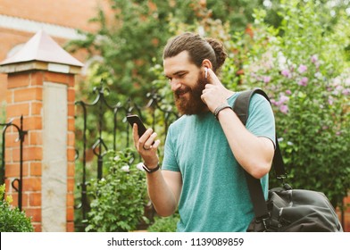 Cheerful happy bearded man looking at his smartphone, listening to the music with earpods outdoors. - Powered by Shutterstock