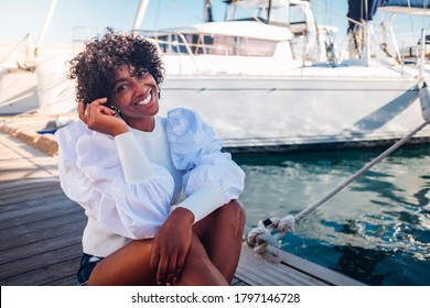 Cheerful happy black african young woman sit down outdor smiling - concept of tourist and summer holiday vacation - boats and dock in background - beauty people african race