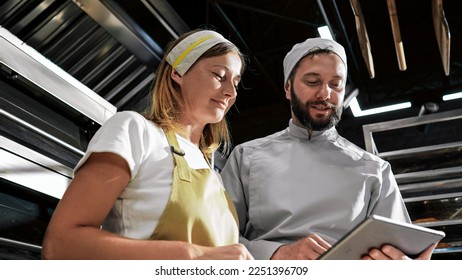 Cheerful happy Caucasian couple of bakers co-workers smiling while watching something on tablet device in bakehouse. Male baker showing something to female colleague on computer. Indoor. - Shutterstock ID 2251396709