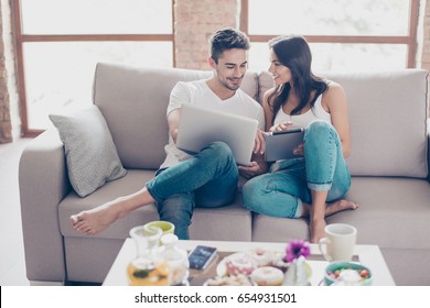 Cheerful Happy Couple Is Doing Online Shopping In Internet At Home Indoors. They Are On Cozy Beige Couch In Casual Clothes, Relaxing And Buying Goods Easily