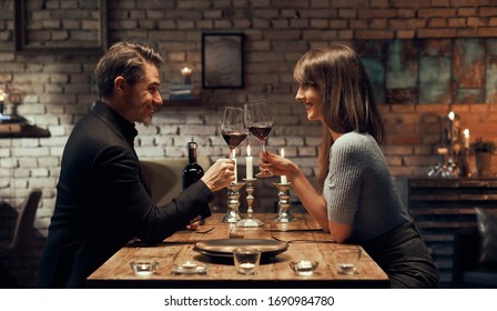 Cheerful Happy Couple Drinking Wine And Having Romantic Dinner At Home. Home Date Night, Clinking Wine Glasses.