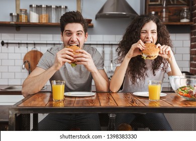 Cheerful Happy Couple Eating Burgers At The Kitchen