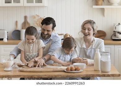 Cheerful happy couple of parents and two cute little kids in aprons teaching to bake, rolling, shaping dough. Children helping mom and dad to prepare dessert, pie, pastry food, - Powered by Shutterstock