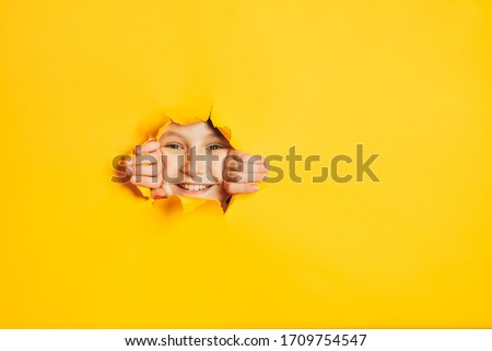 Cheerful happy face of child looking into hole in torn yellow paper wall. Sidebar for text, promotional information, advertising, positive news, events, sale, discount, best choice.