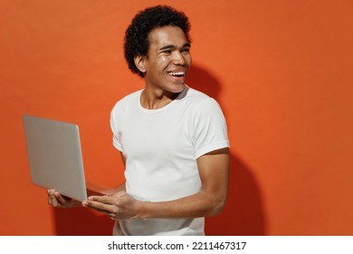 Cheerful Happy Fun Blithesome Young Black Curly Man 20s Years Old Wears White T-shirt Hold Use Work On Laptop Pc Computer Looking Back Behind Isolated On Plain Pastel Orange Background Studio Portrait