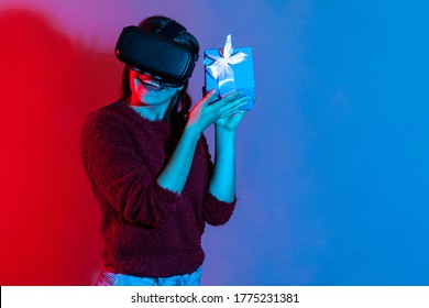 Cheerful Happy Girl Wearing VR Virtual Reality Glasses Holding Gift Box Near Ear, Curious Listening What's Inside Christmas Present, Celebrating Holidays, Birthday. Colorful Neon Light Studio Shot