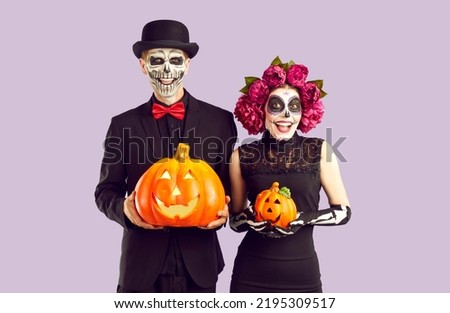 Cheerful happy joyful couple in Halloween costumes with pumpkins in hands on light purple background. They are smiling and looking at camera. Both in black wear with terrible make-ups.