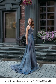 Cheerful Happy Joyful Woman Laughing Merrily On Backdrop Gray Building, Brick Wall, Blooming Flowers. Charming Beauty Smile. Brunette Hairstyle Hollywood Wave. Stylish Elegant Festive Prom Dress Train