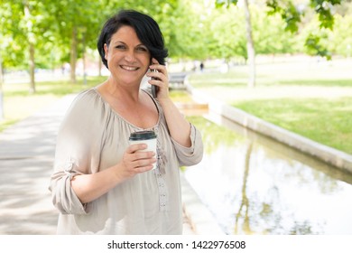 Cheerful Happy Lady Talking On Cell Over Cup Of Coffee In Park. Black Haired Mature Woman Holding Disposable Cup, Speaking On Cellphone And Smiling At Camera. Communication Or Coffee Break Concept