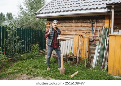 A cheerful and happy man stands proudly in front of a rustic cabin, holding a pile of logs and appears entirely ready to chop wood in a serene and green outdoor environment - Powered by Shutterstock